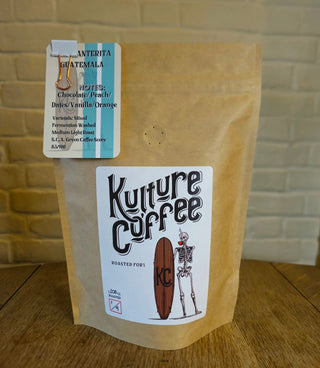 A stand-up kraft paper coffee bag on a wooden table with a light brick wall background. The bag has a large white label reading 'Kulture Coffee' with a skeleton and coffee bean graphic, and a smaller top label indicating Anterita Guatemala coffee.
